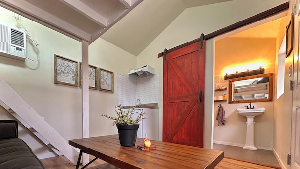 Un espacio de sala acogedor con una mesa de madera, una planta en maceta y una puerta roja que da a un baño.