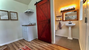 Cocina compacta con refrigerador blanco, estufa y pileta. Una puerta estilo granero roja da a un baño con pileta blanca y un espejo arriba.