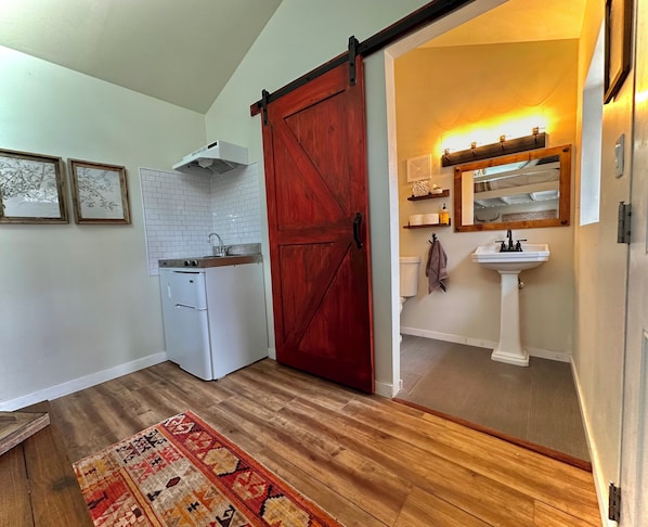 A compact kitchen with a white refrigerator, a stove, and a sink. A red barn-style door leads to a bathroom with a white sink and a mirror above it.