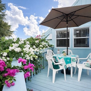 A blue house with a porch, white chairs, a table with a green tablecloth, and a large umbrella. - Seaside Charm: Steps to Beach, Bars, Taxi & Island Fun! (Frigate Bay 78Q7+6Q2 Kittian Village)