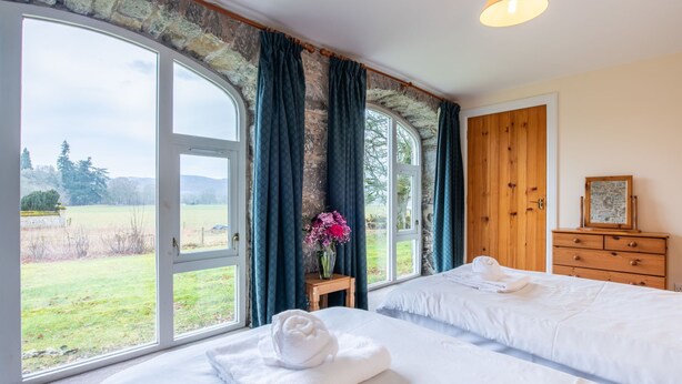 A bedroom with a large window, a wooden dresser, and a bed with white linens. - Old Stables by Interhome (Drumnadrochit)