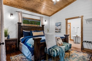 Une chambre avec un plafond en bois, un lit avec une literie bleue, une chaise avec un coussin à motifs et une petite table sur laquelle est posée une plante.