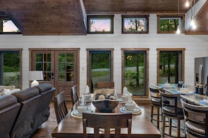 Un coin repas avec une table en bois, des chaises et un bol de collations. Il y a des fenêtres avec des stores et une vue sur la verdure à l’extérieur.