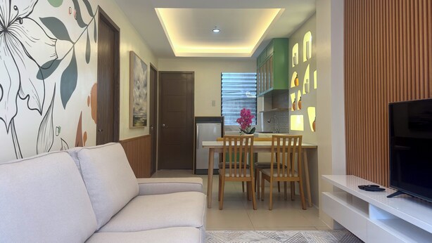 A modern living room with a sofa, a dining table with chairs, a kitchen area with cabinets, and a TV on a stand. - Maple Residences (Talisay)