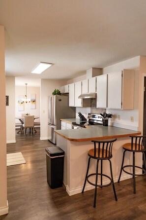 A modern kitchen with a central island, bar stools, and a dining area in the background.