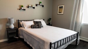 A neatly made bed with a white quilt, a bedside lamp, and a small nightstand with a book. - Family-size condo in the heart of Fairbanks! Short walk to Creamer's field.  (Fairbanks)