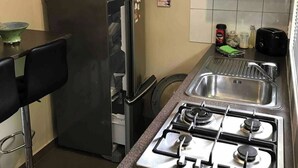 Cocina compacta con estufa a gas, microondas y refrigerador. Hay un lavamanos con grifo, un taburete de bar y un pequeño bote de basura.