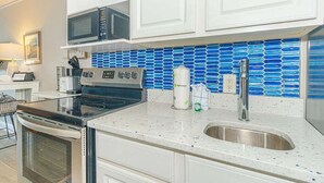 A kitchen with white cabinets, a stainless steel sink, a microwave, a stove, and a blue mosaic backsplash. - Sea Mist 51108 (Myrtle Beach)