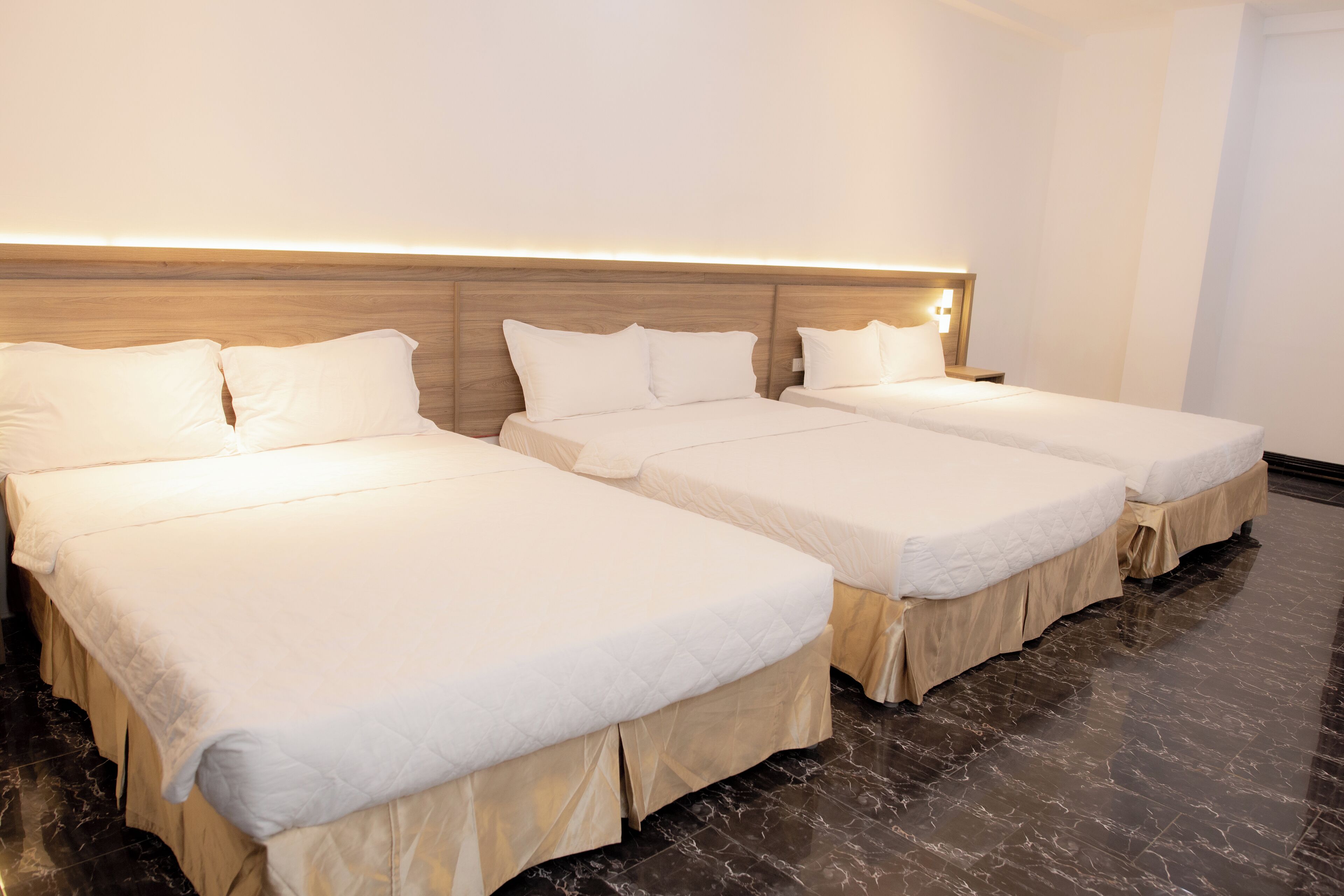 Une chambre d’hôtel avec trois lits simples, une tête de lit en bois et un sol en marbre.