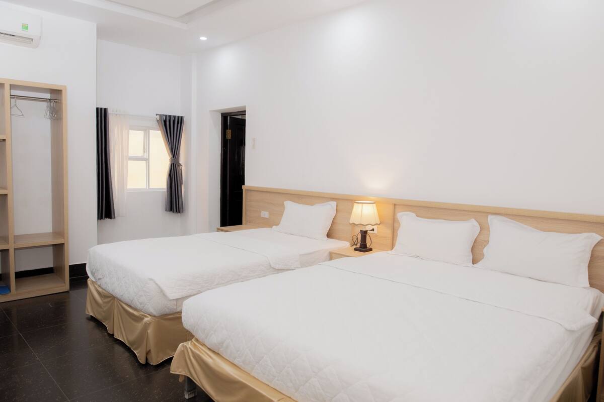 A hotel room with two beds, a wardrobe, a window with curtains, and a nightstand with a lamp.