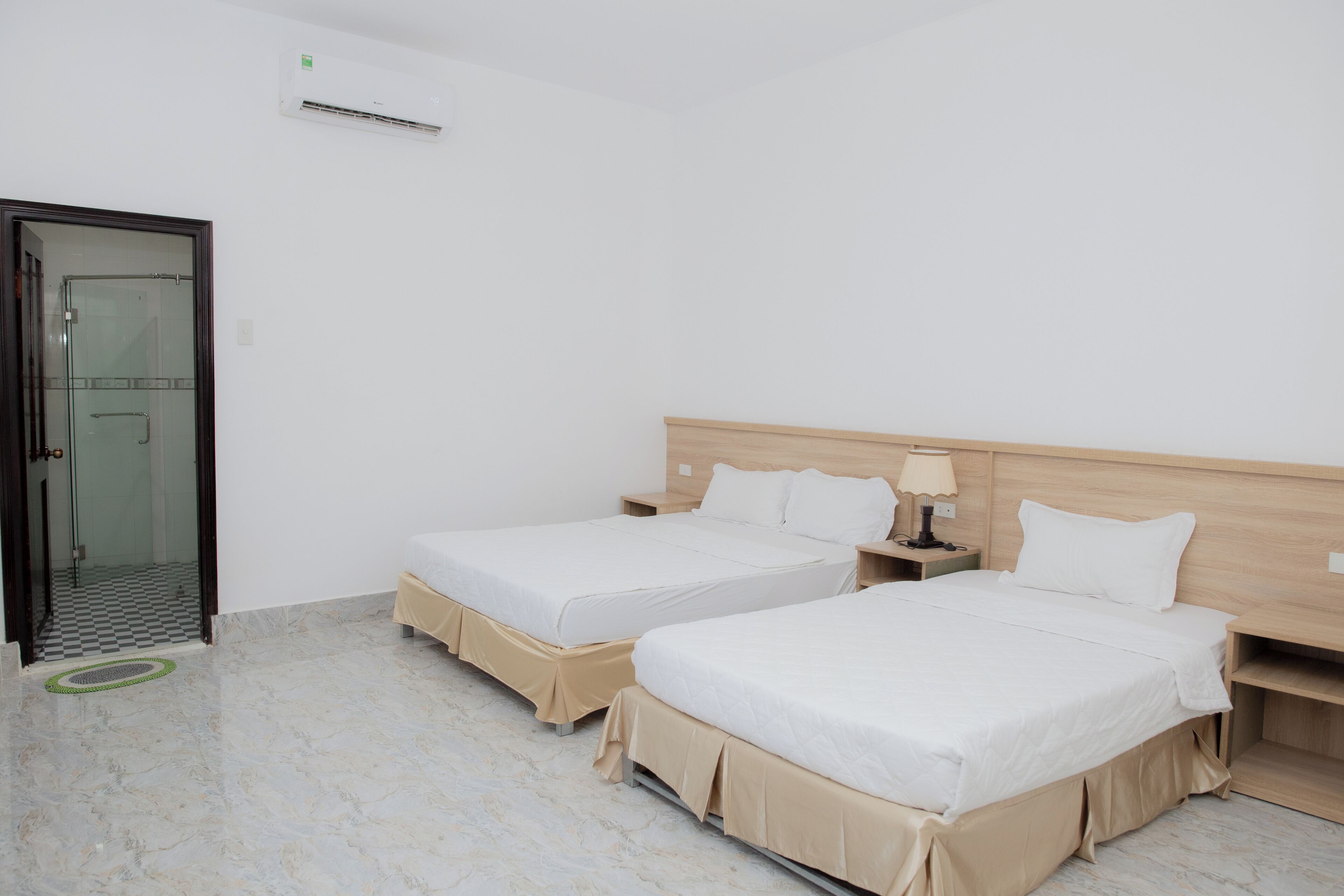 Habitación de hotel con dos camas, una mesita de noche pequeña con lámpara, cabecero de madera y puerta de vidrio que da al baño.