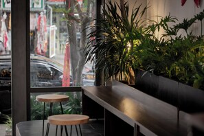 A modern interior with a wooden bench and two stools, a large potted plant, and a view of the street through a glass window. - Mien Tay Hotel (Can Tho)