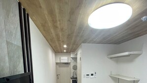 A narrow hotel room with a bed, a TV, a small kitchenette, and a ceiling light. - JR/ (Shinjuku Ku Okubo)