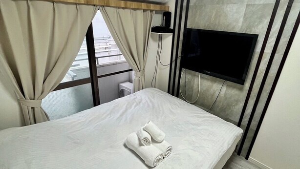 A modern hotel room with a bed, curtains, an air conditioning unit, a TV, and a window with a view. - JR/ (Shinjuku Ku Okubo)