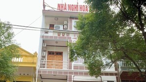 Un immeuble à plusieurs étages, sur lequel on peut lire l’inscription « NHÀ NGHÌ HOA SỮA », ce qui indique qu’il s’agit d’un hôtel. Des plantes en pot sont disposées devant l’entrée, et une personne se tient devant l’immeuble.