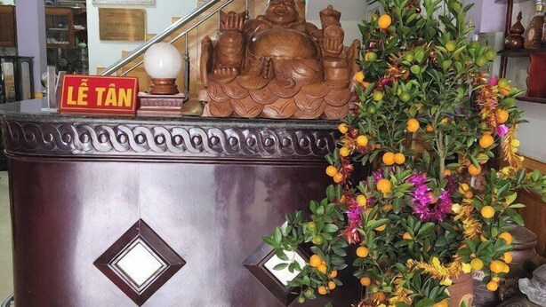 Un comptoir d’accueil orné d’une statue de Bouddha décorative, une enseigne indiquant « LE TAN » et un pot de fleurs avec des fleurs oranges.