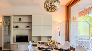 A modern dining area with a wooden table set for four, surrounded by white chairs. A television is mounted on the wall, and there are shelves with various items above it. - Attilio lake front view by Interhome (Ispra)