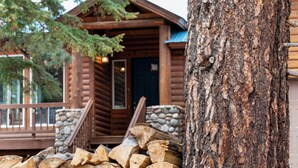 A log cabin with a stone foundation, a wooden deck, and a stack of firewood.