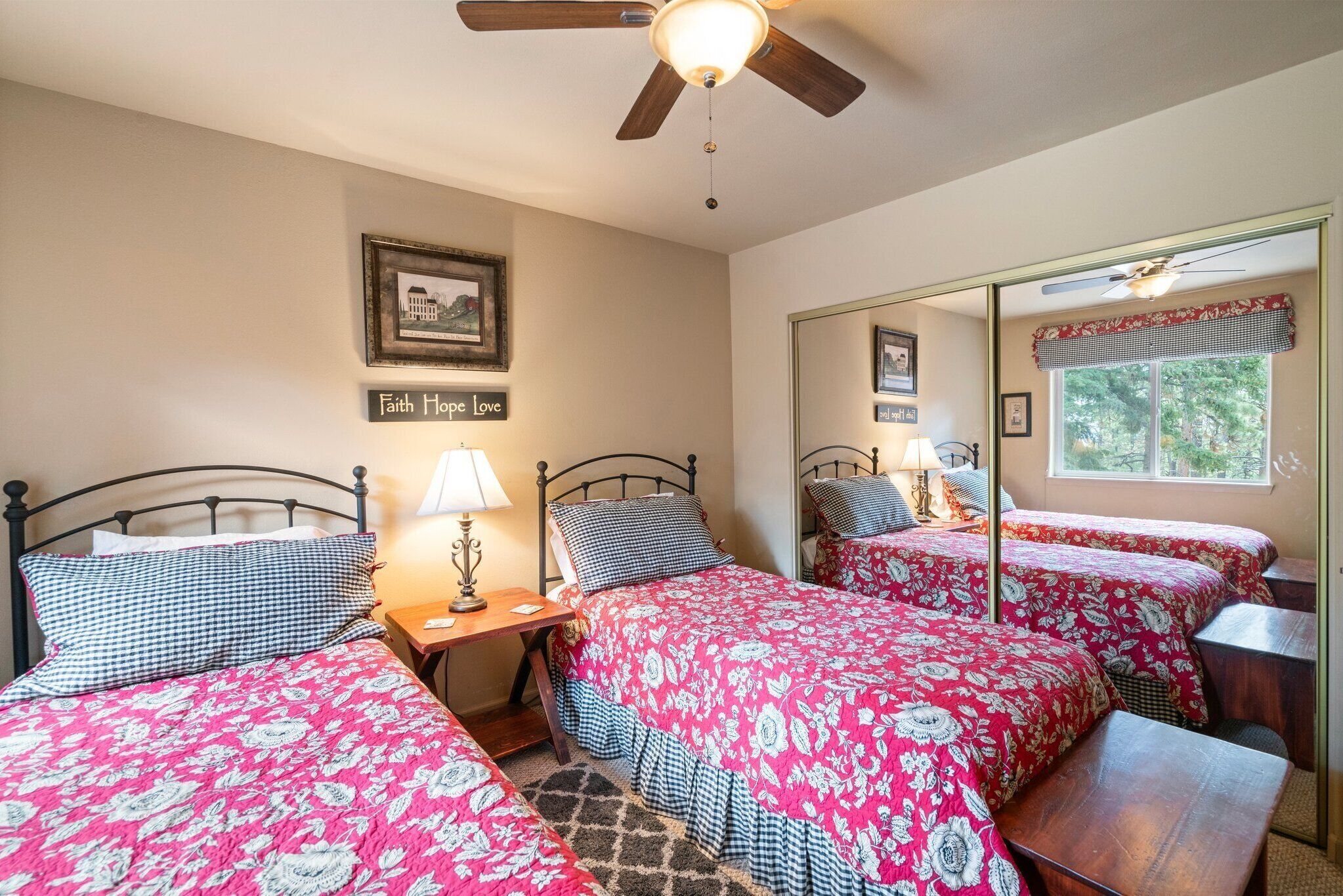 A bedroom with two beds, a ceiling fan, a mirror, and a window with a view of trees.