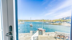 Vista costera desde una ventana que da a un puerto con barcos, un muro de piedra y edificios al fondo.