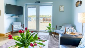Una sala de estar con televisor, dos sillones, una mesa con un vaso, y vista a la playa a través de un ventanal.