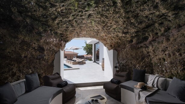 Un interior moderno con una zona de estar circular, una mesita con una revista y vistas a una piscina exterior a través de un arco natural de roca.