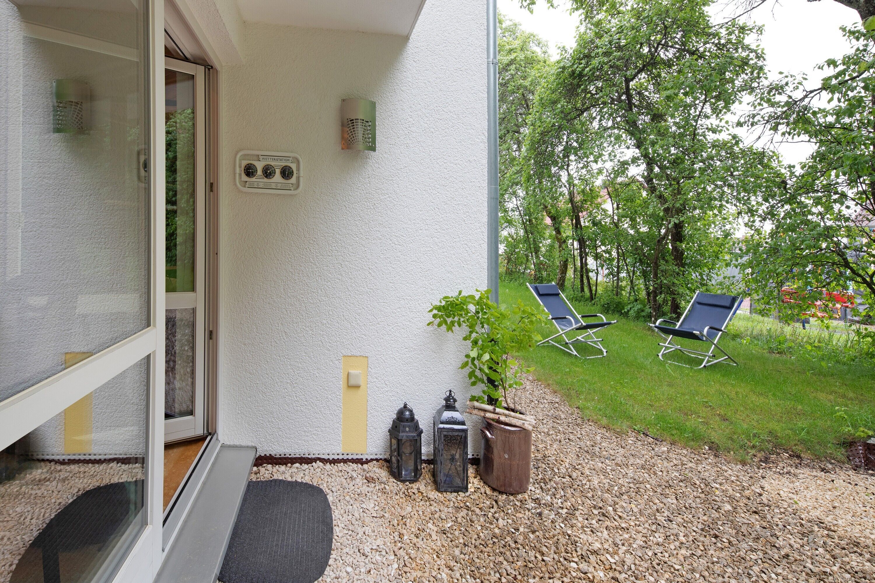 En balkong med en glasdörr, en liten krukväxt, två lyktor och två trädgårdsstolar.