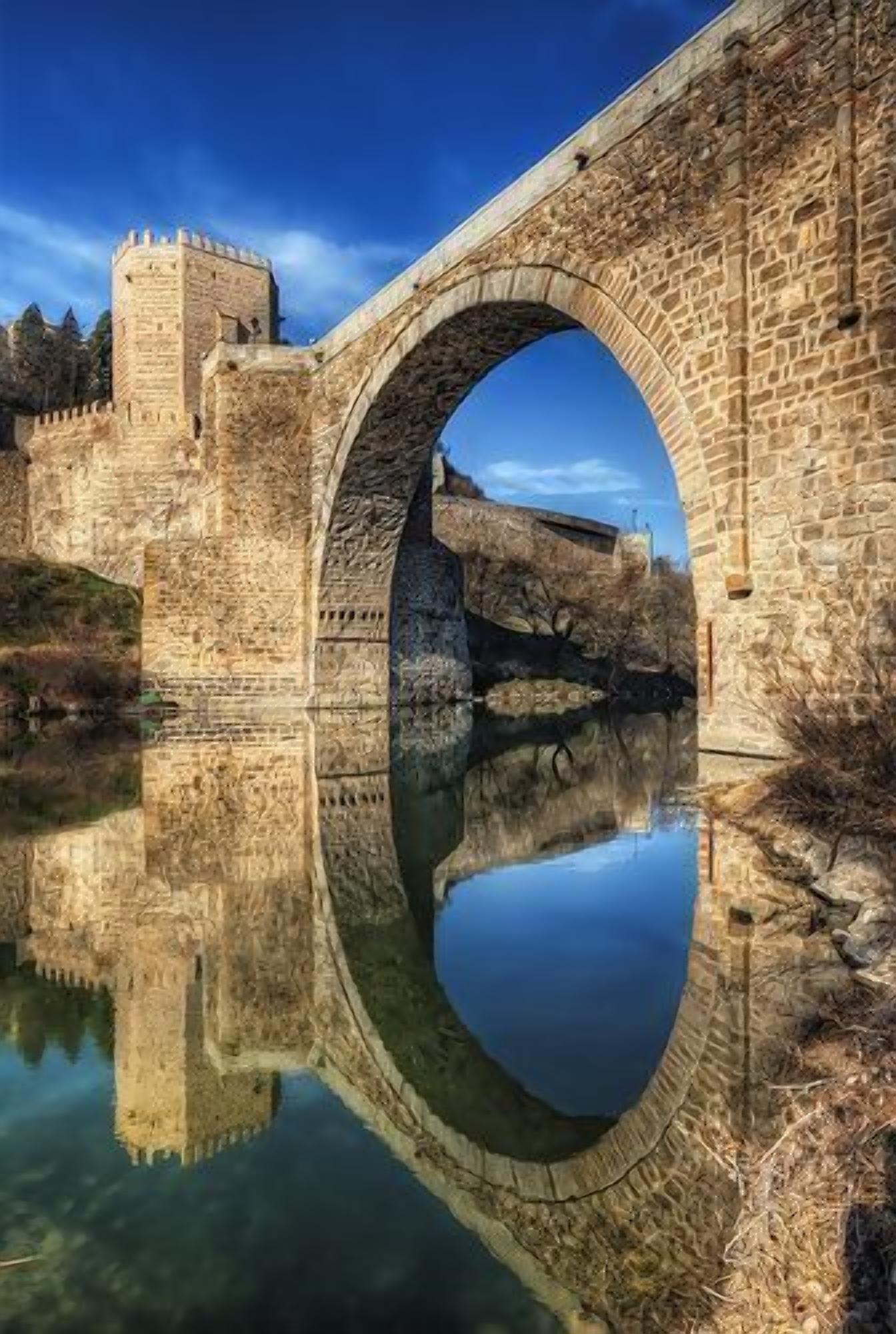 Un puente de piedra con un reflejo claro en el agua, una estructura similar a un castillo a la izquierda y un cielo azul despejado.