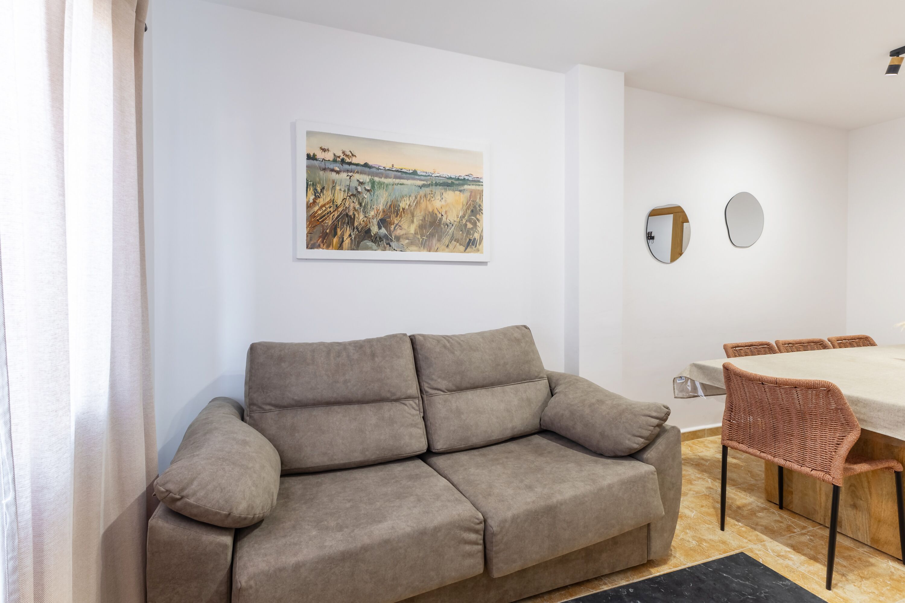 Una sala de estar con sofá, una mesa de comedor con sillas, un cuadro en la pared y un espejo.