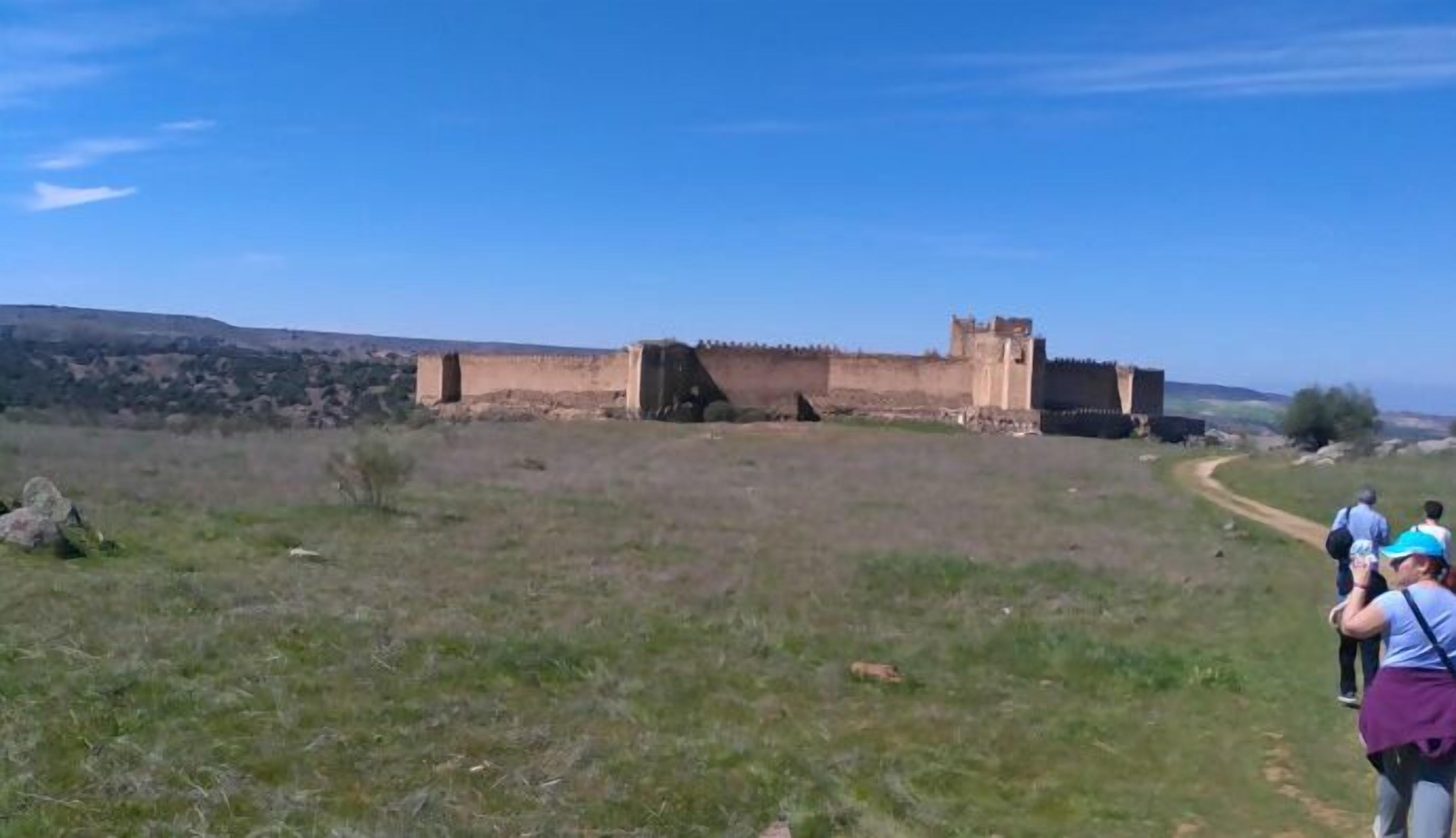 A historical fortification with a clear view of its walls and towers, situated in a grassy field with a dirt path leading to it.