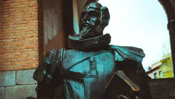 Une statue d’une figure historique, vêtue d’une tenue formelle, est placée devant un mur de briques et une porte en arc de cercle.