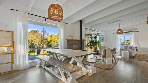 A spacious room with a large wooden table, white curtains, and a view of trees through large windows. - Lake Metung | Modern Lake House Escape (Metung)