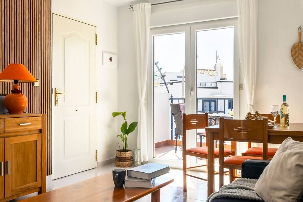 Una sala de estar acogedora con un mueble de madera, una mesa con libros y una lámpara, una planta en maceta y vistas al balcón exterior.