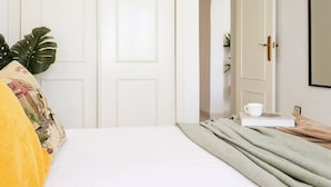 Een net opgemaakt bed met een witte deken, een groene plaid en een geel kussen. Op het bed staan een witte beker en een boek.