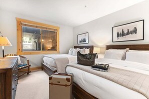 Une chambre d’hôtel avec deux lits, un bureau en bois, une chaise, une valise et des tableaux encadrés aux murs.