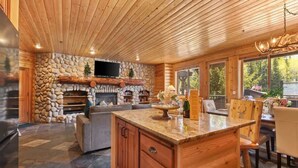 A spacious living area with a stone fireplace, wooden ceiling, and a large window overlooking a forested area.