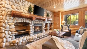 A log cabin interior with a stone fireplace, wooden ceiling, and a cozy seating area.