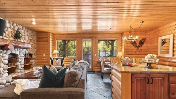 A log cabin interior with a stone fireplace, a sofa, a dining area, and a kitchen with wooden cabinets.