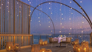 An outdoor dining area with a table set for two, surrounded by wicker chairs and candlelit lanterns. - Centara Mirage Lagoon Maldives (Kodhipparu Falhu)