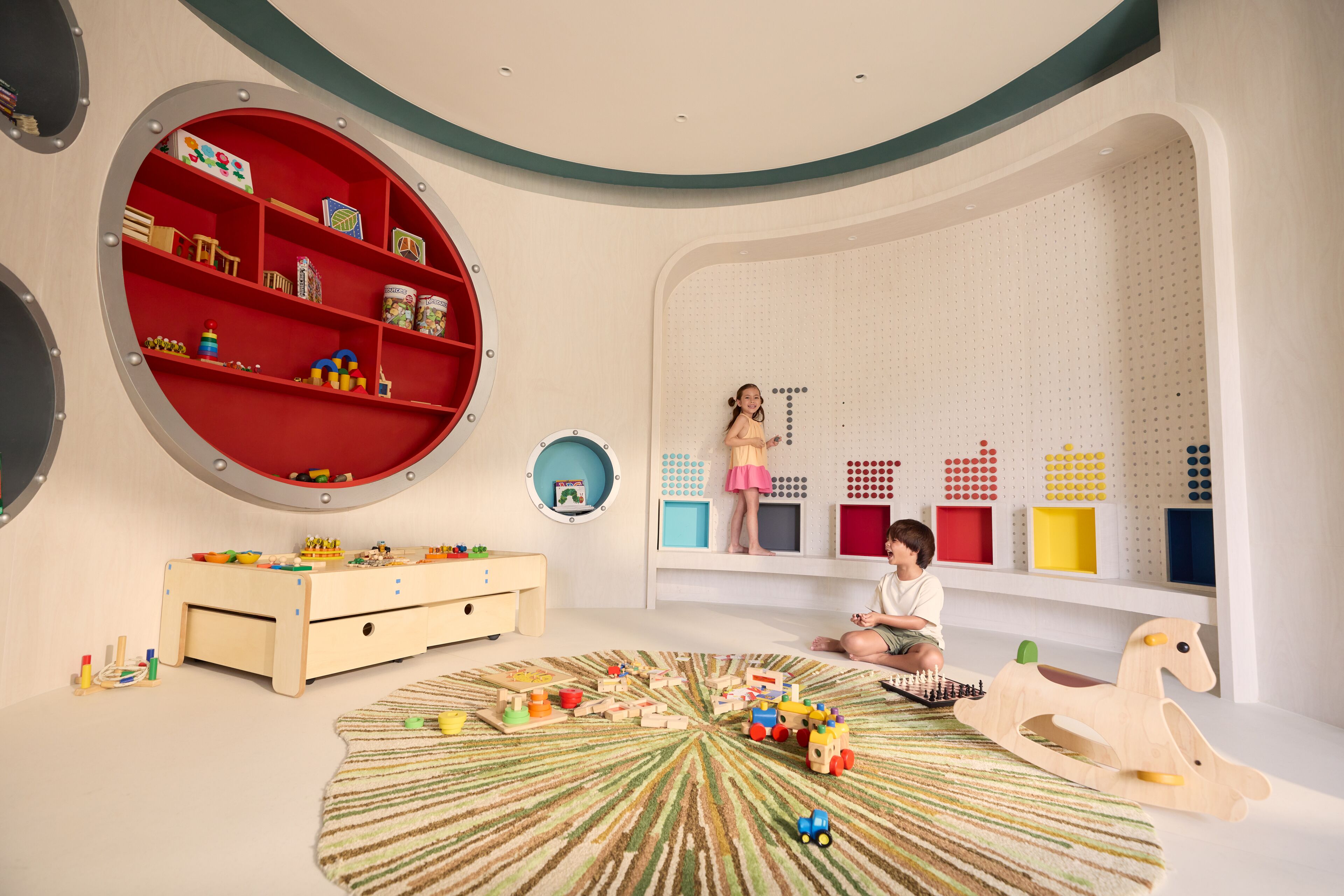 A child playing with toys in a room with a large red circular shelf, a wooden play table, and a colorful play mat.