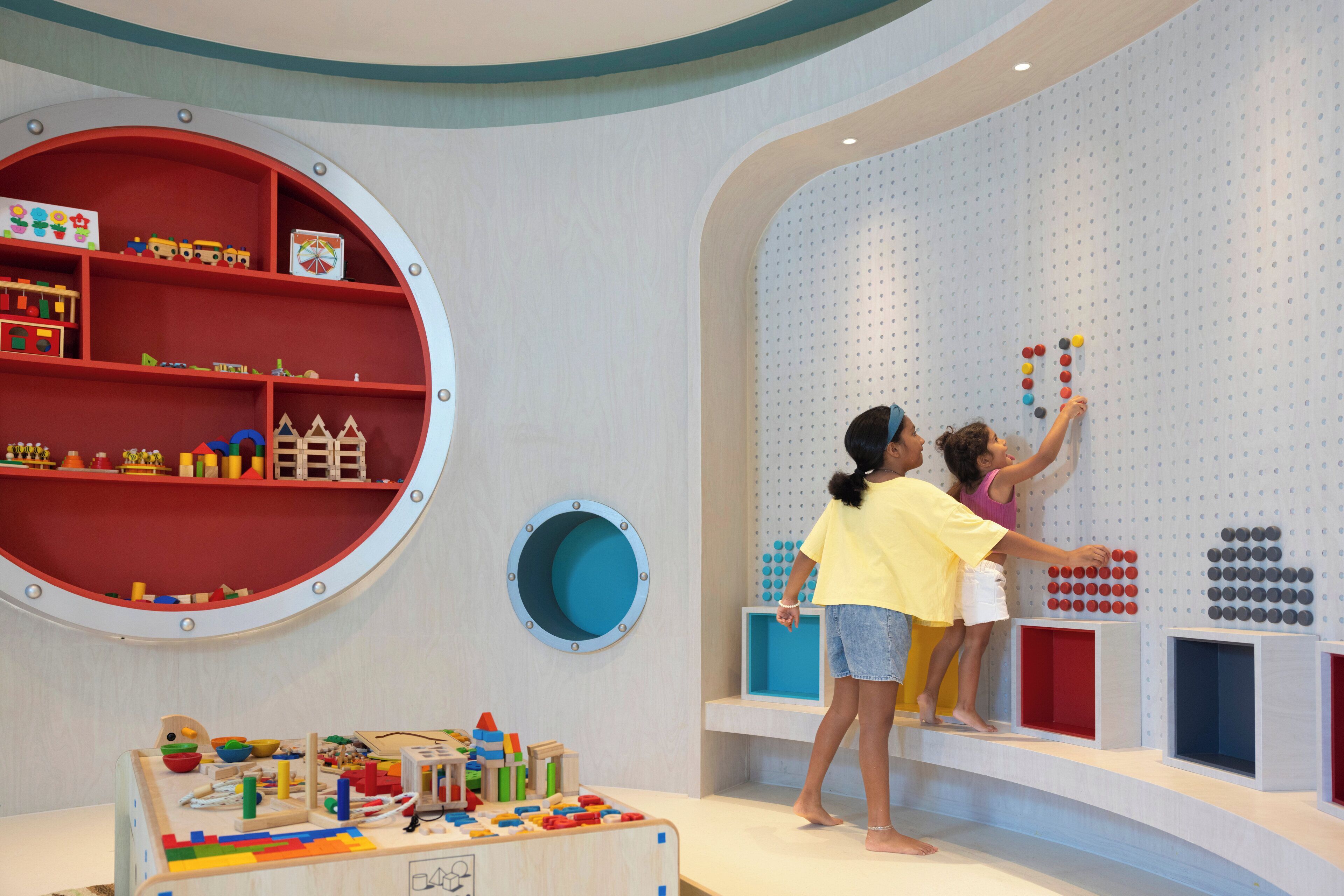 Zona de juegos con un estante circular lleno de juguetes, una ventana circular azul y un panel de usos múltiples para colgar juguetes.
