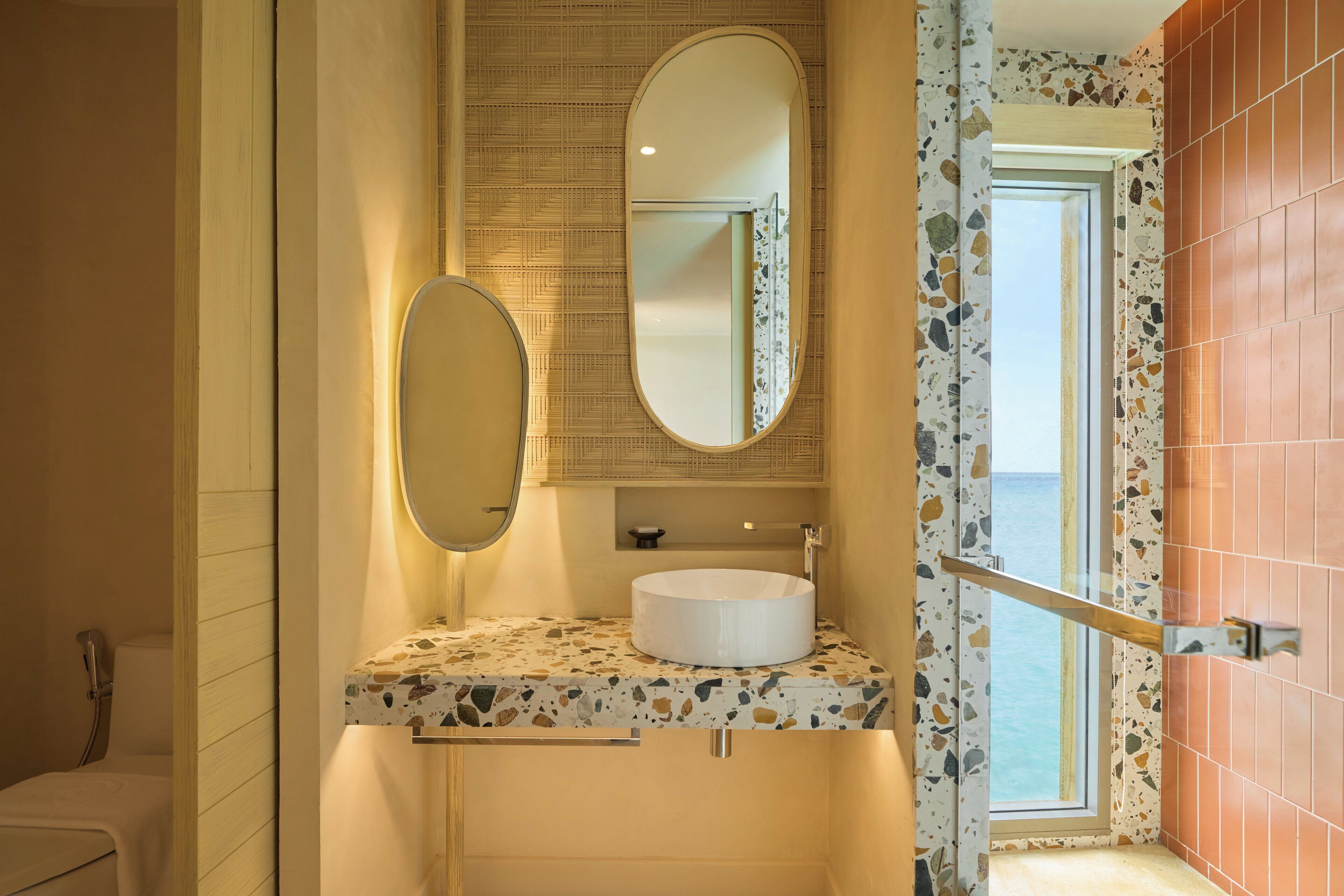 A bathroom with a round sink, two mirrors, and a view of the sea through a window.
