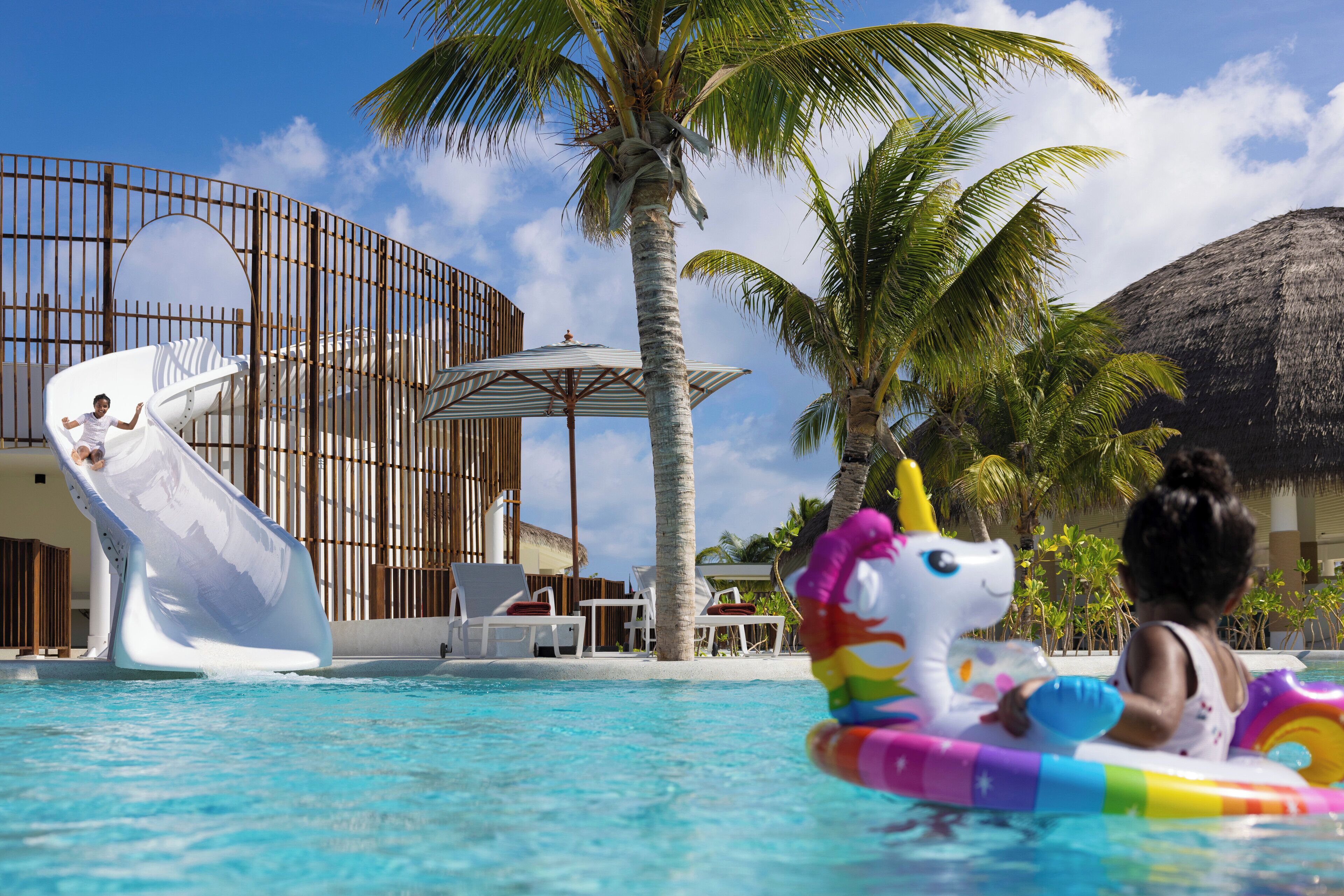 Un espace piscine doté d’un toboggan, de chaises longues et de palmiers.