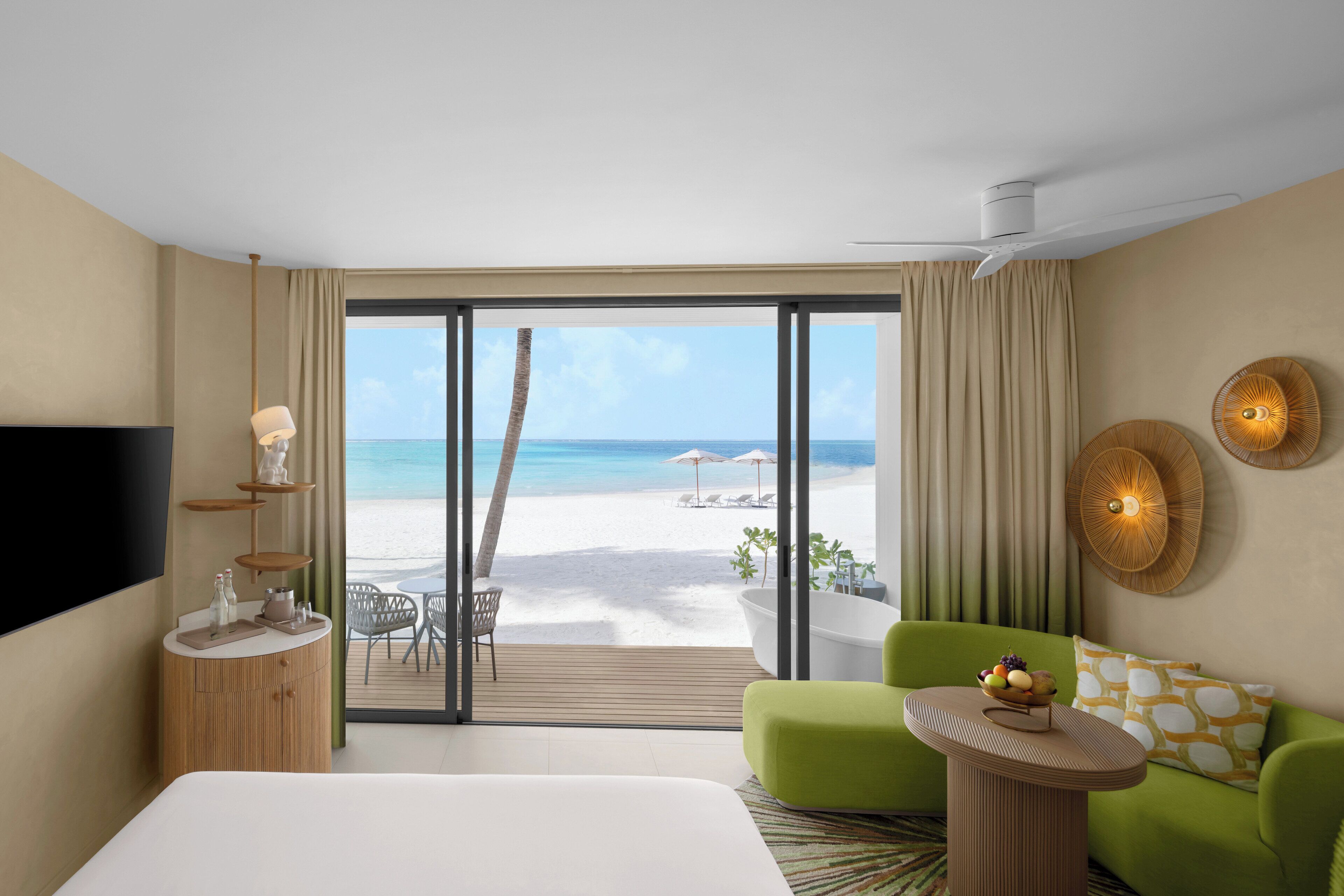 A hotel room with a large bed, a green armchair, a small table with a fruit bowl, a TV, a wooden side table, a glass-top table, and a view of the beach.