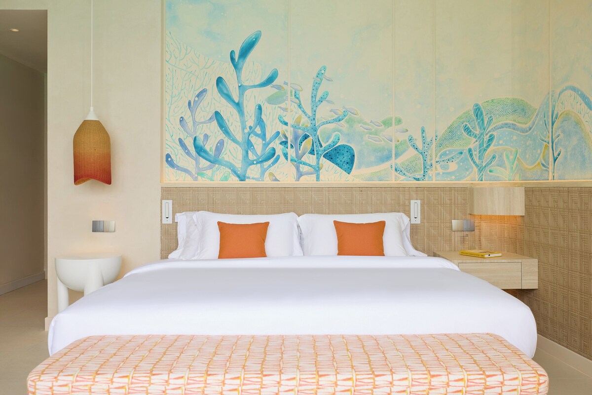 Ein Hotelzimmer mit einem großen Bett, weißer Bettwäsche, orangefarbenen Kissen, einer Wandmalerei von Unterwasserpflanzen, einer Hängeleuchte und einem Nachttisch mit einer Lampe.