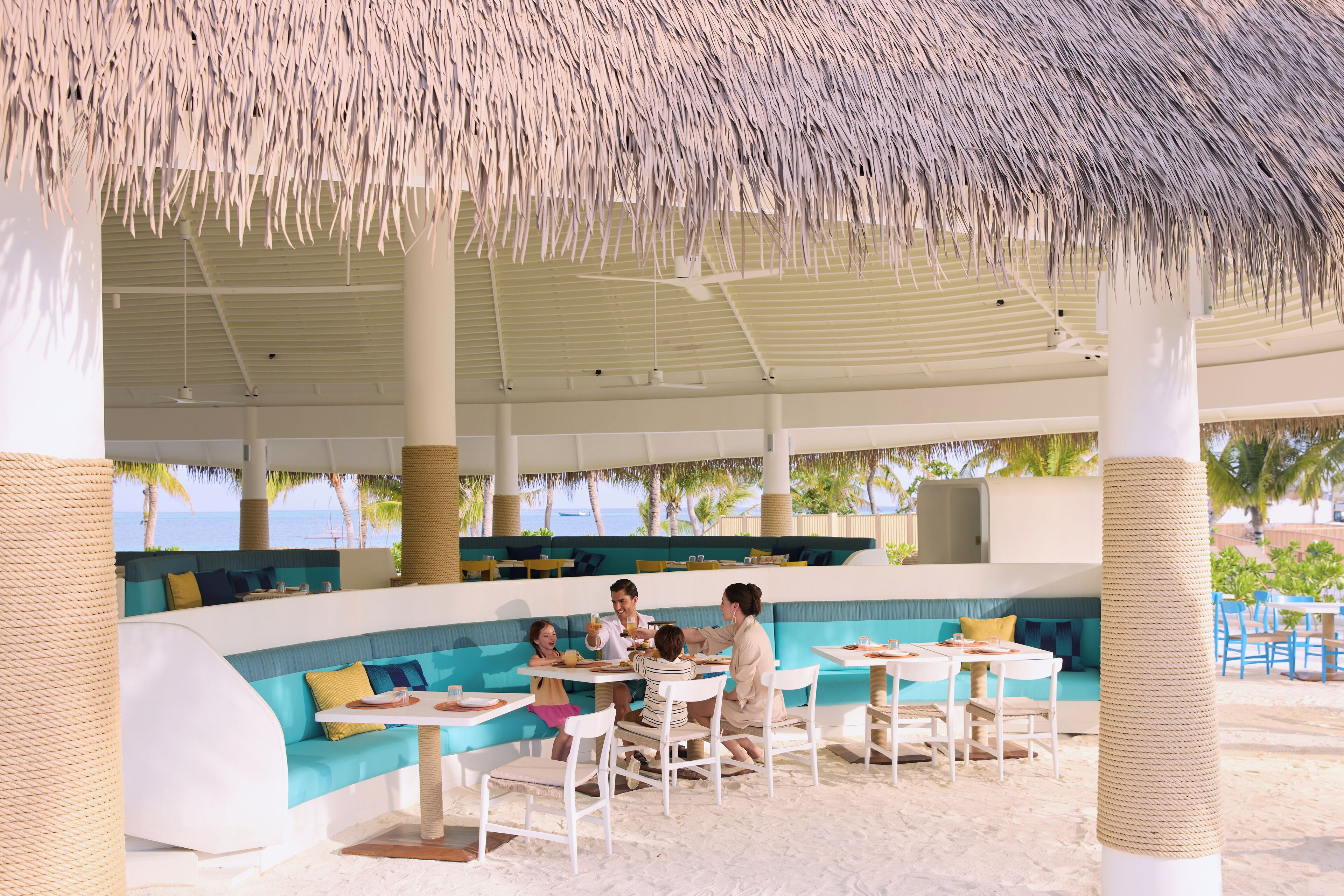 A beachside restaurant with outdoor seating, a thatched roof, and a view of the ocean.