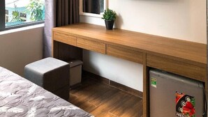 A hotel room with a wooden desk, a television, a small refrigerator, a window with curtains, and a plant.