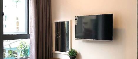 A hotel room with a wooden desk, a television, a small refrigerator, a window with curtains, and a plant.