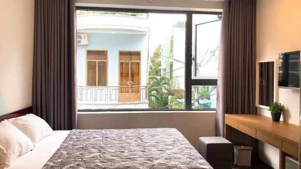 A hotel room with a bed, a desk, a TV, and a window with a view of a building and greenery.