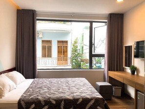 Une chambre d’hôtel équipée d’un lit, d’un bureau, d’une télévision et d’une fenêtre donnant sur un immeuble et de la verdure.